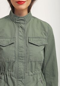 Oliwkowa kurtka robocza wykonana z tkaniny bawełnianej, z wysokim kołnierzem, dwiema kieszeniami na piersi, przednim zamkiem błyskawicznym oraz detalami w talii.