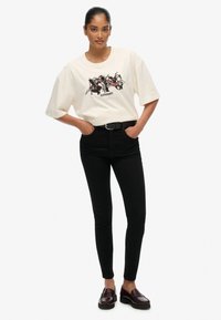 T-shirt oversize color avorio con stampa grafica, abbinato a jeans neri a vita alta e mocassini borgogna scuri. Outfit semplice e casual.