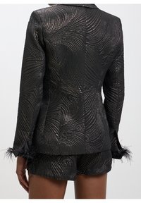 Schwarzer Blazer mit einem strukturierten Wirbelmuster, metallischen Fäden und gefiederten Manschetten. Kombiniert mit passenden schwarzen Shorts, die ein figurbetontes Design aufweisen.