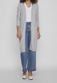 Ljust grå lång kofta med ribbade manschetter och fickor, bärs över en vit V-ringad t-shirt, ihop med vida blå jeans och krämfärgade skor.