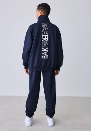 Chłopiec stoi tyłem do kamery, ubrany w granatowy dres z pionowym napisem „BAKER” na kurtce oraz białe trampki.