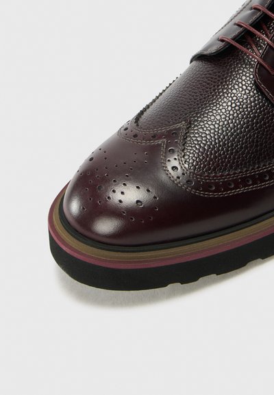 Scarpa brogue in pelle bordeaux con tomaia texturizzata, dettagli perforati e suola a strisce multicolori. Punta arrotondata e design con lacci.