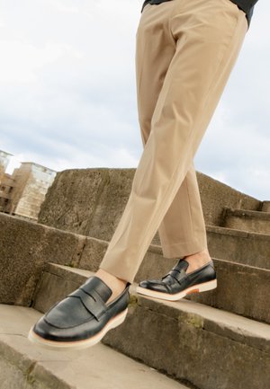 Persona con pantaloni beige e mocassini in pelle nera che scende scalini di pietra all'aperto.