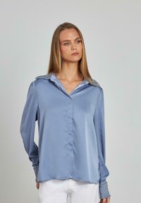 Blusa de satén azul claro con cuello que presenta tachuelas plateadas, mangas largas con puños adornados con tachuelas y una superficie lisa. Cierre delantero de botones.
