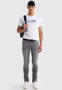 Giovane uomo con cappellino nero, maglietta bianca con scritta "BIG STAR", jeans grigi, sneakers bianche e zaino nero su una spalla, in piedi dritto.