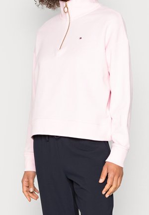 Persona llevando una sudadera ligera color rosa claro con cremallera hasta el pecho y un pequeño logo en el pecho, pantalones negros, de pie frente a un fondo liso.