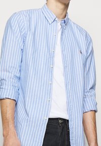 Jasnoniebieska koszula w paski z kołnierzykiem zapinanym na guziki, z białymi pionowymi paskami, miękkim materiałem i małym haftowanym logo na piersi.