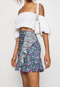 Jupe portefeuille florale avec un ourlet volant, principalement bleu foncé avec des fleurs roses et blanches. Associée à un haut smocké blanc court et un sac texturé.