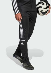 Czarne spodenki sportowe z białym wzorem w trzy paski. Materiał jest gładki i dopasowany. Czarne buty z teksturowanym wykończeniem i ostrymi korkami.