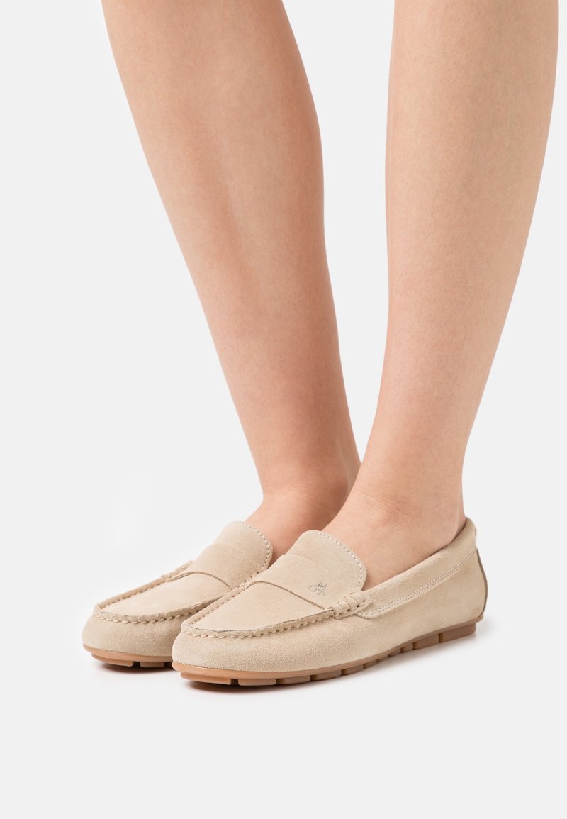 Mocassins en daim beige avec semelles en caoutchouc portés pieds nus, montrant des angles latéraux et avant sur un fond clair uni.