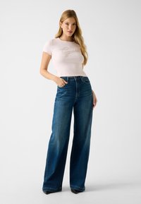 Light pink cropped t-shirt with "Guess" logo, paired with high-waisted, wide-leg blue denim jeans. Smooth texture and classic design.