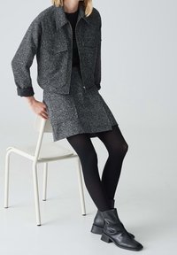 Femme assise sur une chaise blanche portant une veste grise texturée, une jupe assortie, des collants noirs et des bottines noires sur fond uni.