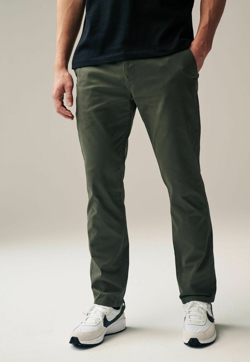 Olive green chinos made of cotton blend, featuring a slim fit, front pockets, and back pockets, paired with white and black athletic shoes.
