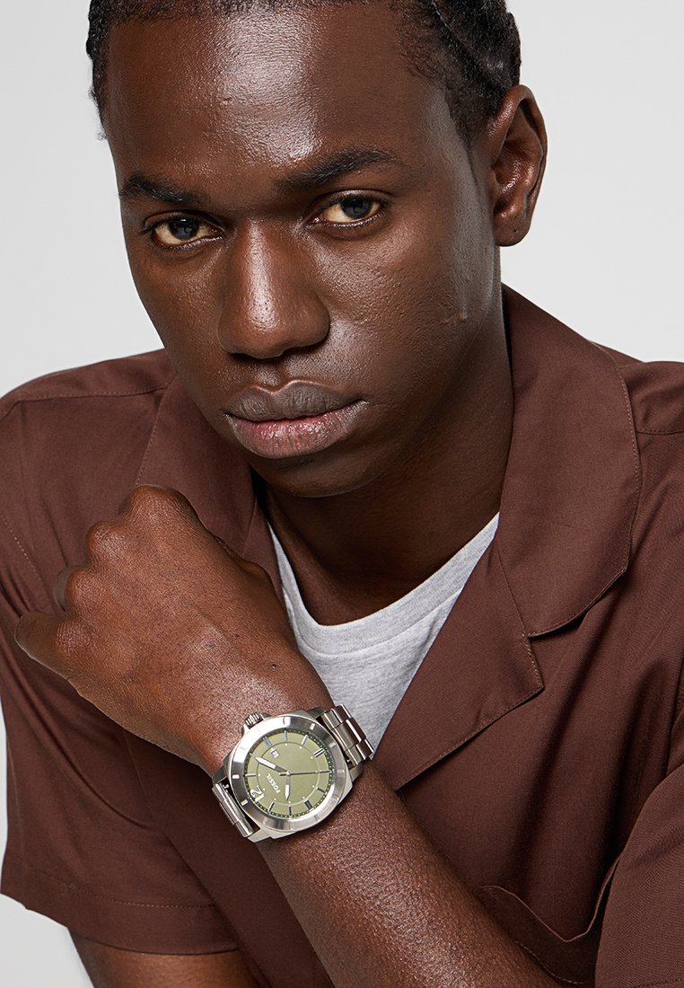 Orologio in acciaio inossidabile argentato con quadrante verde, dotato di indicatori delle ore e di un robusto bracciale, indossato su un polso con abbigliamento marrone.