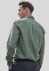 Camisa verde de manga larga hecha de tejido ligero, con cuello clásico, puños abotonados y un pliegue en la parte posterior para mayor facilidad de movimiento.