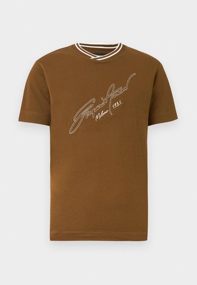 Καφέ κοντομάνικο T-shirt με λευκή ριγέ V-λαιμόκοψη και λευκό χειρόγραφο κείμενο «Giorgio Armani Milano 1981» στο στήθος.