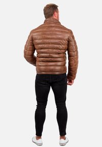 Giacca puffy in pelle marrone con colletto alto e motivo di cuciture orizzontali, indossata con jeans neri aderenti e sneakers bianche.