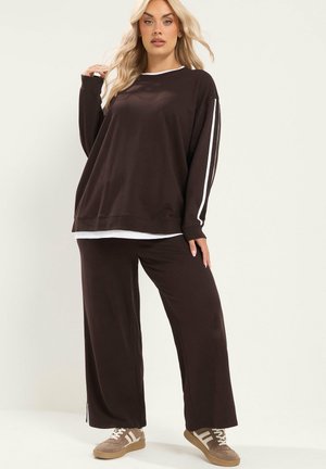Vrouw met lang blond haar die een donkerbruine losse trui en wijde broek draagt met beige sneakers tegen een effen lichte achtergrond.