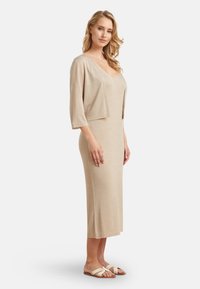 Beige, knee-length dress with a V-neck and short sleeves; made of soft, textured fabric; paired with flat, open-toed sandals.