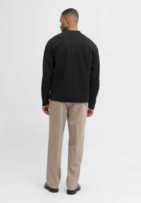 Férfi fekete hosszúujjú pólót, bézs nadrágot és fekete cipőt viselve áll, háttal a fényes fehér háttérnek.