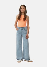 Pomarańczowy bezrękawnik w połączeniu z jasnoniebieskimi spodniami o szerokich nogawkach. Spodnie mają kieszenie i postrzępiony dół. Białe sneakersy dopełniają stylizację.