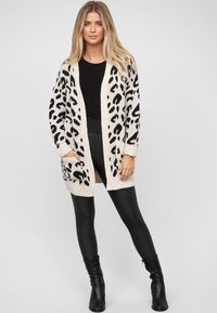 Lichtgewicht beige cardigan met zwart luipaardprint. Open voorkant, lange mouwen en twee voorzakken. Gecombineerd met een zwarte top en leggings.