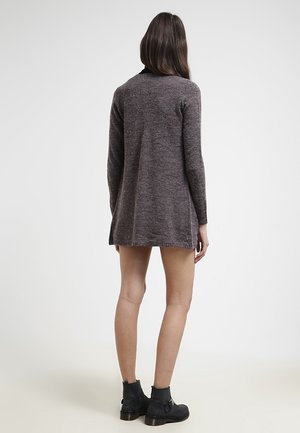 Femme aux longs cheveux foncés, de dos, portant une robe-pull grise à manches longues et des bottines noires sur un fond clair.