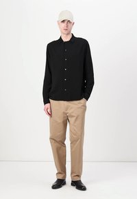 Schwarzes langärmliges Hemd mit Knöpfen, beige Hose und schwarze Schuhe; Modell trägt eine hellbeige Kappe und steht vor einem schlichten weißen Hintergrund.