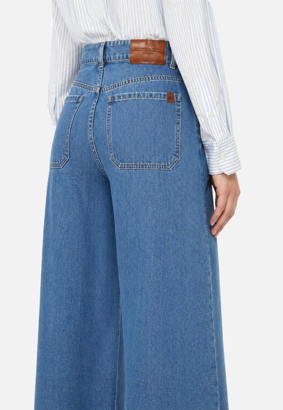 Jeans blu a vita alta e gamba larga con tasche posteriori e patch in pelle marrone del marchio, indossati con una camicia bianca a righe blu a manica lunga.