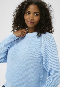 Femme avec des cheveux bouclés portant un pull en maille bleu clair, tenant le col d'une main, regardant légèrement sur le côté.
