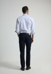 Camisa de botones a rayas azul claro, jeans azul oscuro con detalles de costura y zapatos marrones. Se muestra la vista trasera del atuendo.