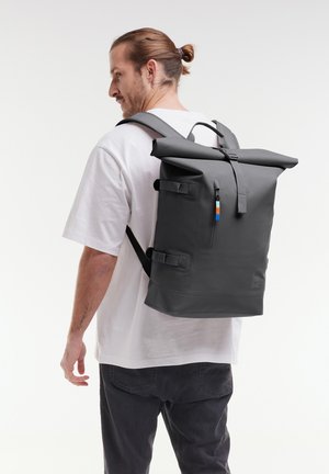 Sac à dos gris à rabat, fabriqué en matériau durable, avec des sangles réglables, une poche zippée à l'avant et un détail coloré sur la tirette.