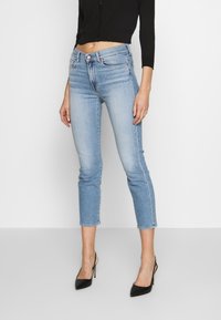 Jeans recortados azul claro con un ajuste slim, cinco bolsillos y cierre de botón, combinados con tacones de aguja negros. Textura de denim suave.