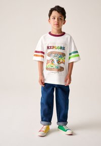 Fehér póló „EXPLORE” grafikával és színes sportcipő ábrázolással. Bordó, narancssárga és zöld csíkok a vállakon. Kék farmernadrág.