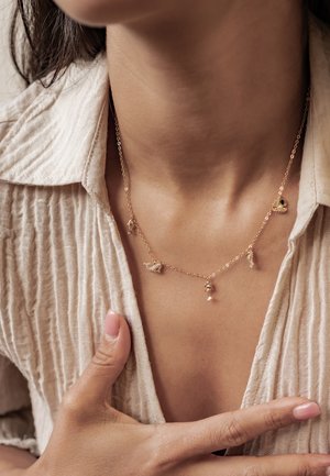 Nahaufnahme einer Person, die ein beigefarbenes Hemd und eine filigrane goldene Halskette mit kleinen anhängerförmigen Tiermotiven trägt.