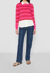 Maglione a righe fucsia e rosso con colletto bianco, indossato sopra una camicia bianca. Abbinato a jeans di denim blu a vita alta e sandali beige.