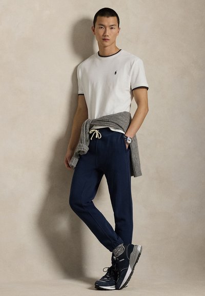 Muž stojaci pri béžovej stene, oblečený v bielom tričku s krátkym rukávom, námorníckych teplákoch, sivom svetri zviazanom v páse a čiernych teniskách so sivými ponožkami.