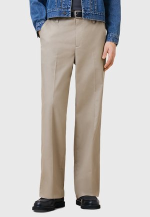 Beige lige benbukser med en glat tekstur, der har sidelommer og subtile frontfolder, parret med sorte sko.