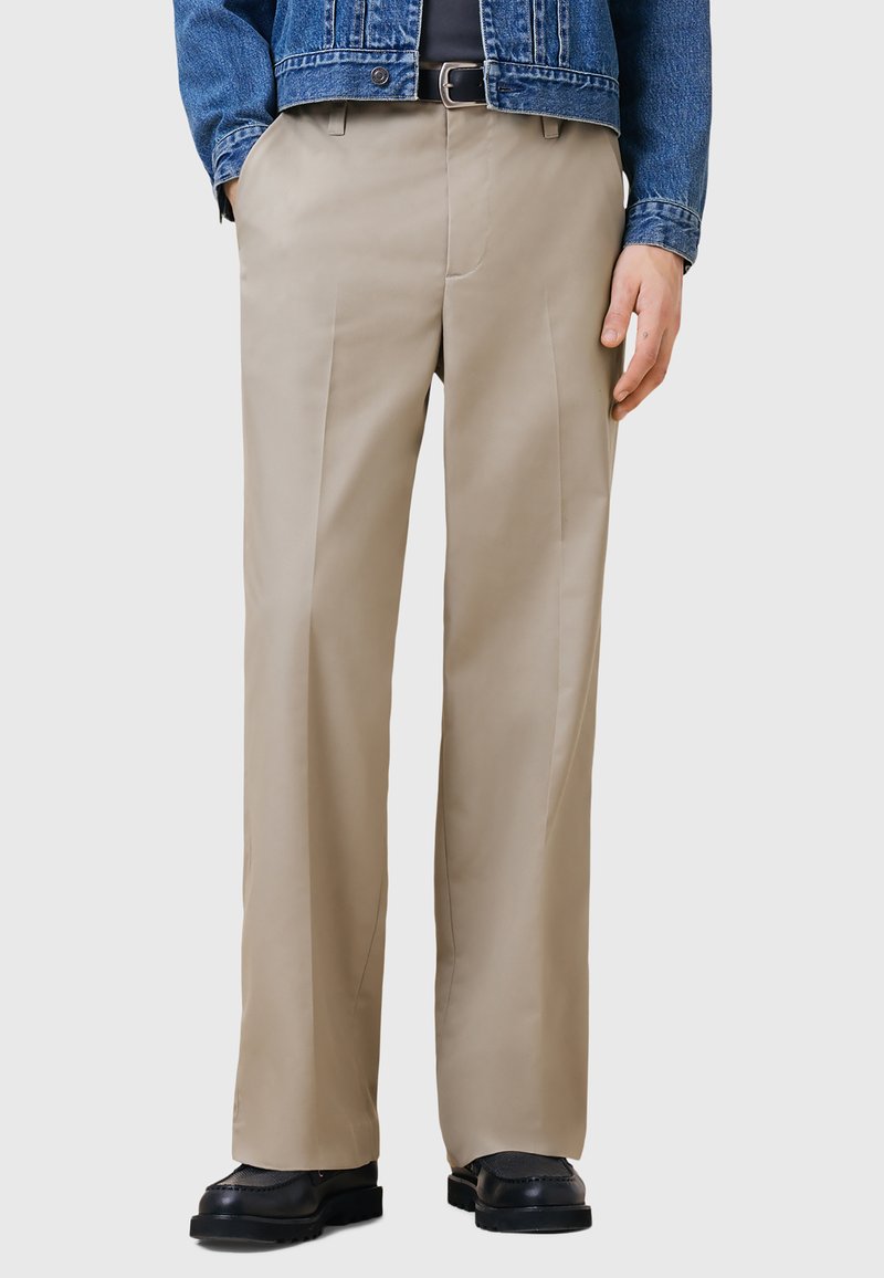 Beige rechte broek met een gladde textuur, voorzien van zijzakken en subtiele voorkreukels, gecombineerd met zwarte schoenen.