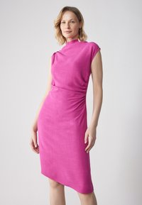Robe rose ajustée avec un col haut et des manches courtes. Présente des détails froncés à la taille et une ourlet asymétrique. Texture lisse.