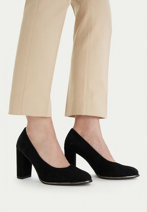 Pies calzados con zapatos de tacón grueso de ante negro combinados con pantalones beige recortados sobre un fondo claro y sencillo.