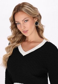 Schwarzer Pullover mit einer weißen gerippten V-Ausschnitt, ausgestattet mit einer strukturierten Strickerei und einer lockeren Passform. Goldene und schwarze auffällige Ohrringe runden das Outfit ab.