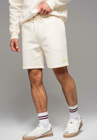 Krémové fleecové šortky s elastickým pasem, bočními kapsami a malou zelenou nášivkou. Vytvořeno s bílými teniskami a pruhovanými ponožkami.