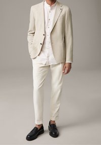 Uomo che indossa un blazer beige, una camicia bianca con bottoni, pantaloni crema e mocassini neri, in piedi contro uno sfondo neutro e semplice.