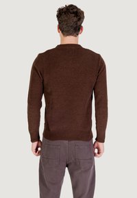 Maglione lavorato a maglia marrone con una finitura testurizzata, polsini e orlo a coste, design aderente; indossato con pantaloni grigi, visto da dietro.
