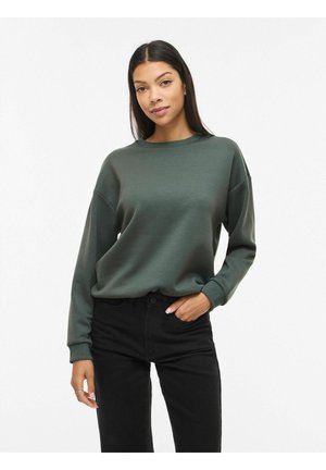 Pikkade tumedate juustega naine, kes kannab tumerohelist dressipluusi ja musti pükse, seisab ühe käega taskus valge tausta ees.