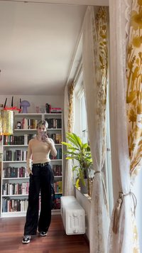 Beige ribbed pitkähihaine paita, mustat leveälahkeiset housut vyöllä, sisätila, jossa on kirjahyllyjä, verhoja ja vihreä kasvi.