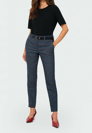 Mujer con top negro de manga corta, pantalones ajustados de cuadritos con cinturón negro y tacones rojos de punta, de pie con una mano en el bolsillo.