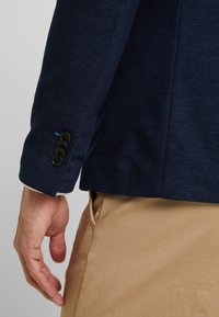 Marinblå blazer i ett texturerat tyg, med en fyra-knapps manschett och en diskret blå accenttråd nära fållen. Beiga byxor syns.