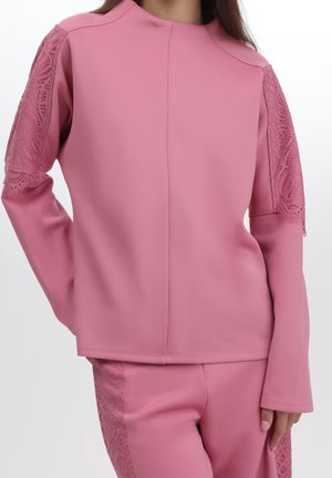 Vrouw draagt een roze top met lange mouwen en bijpassende broek met kanten details aan de mouwen en zijkanten van de broek, tegen een witte achtergrond.